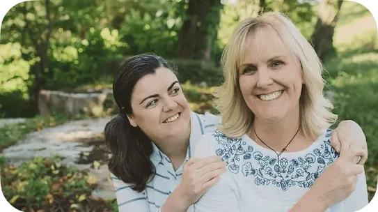 A younger woman with dark hair tied back in a ponytail looks affectionately at a smiling older blonde woman. The blonde woman smiles directly at the camera while the younger woman embraces her from behind in a lush, green outdoor setting.