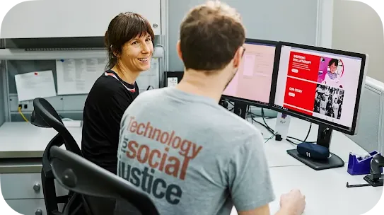 A woman sitting at a desk cubicle smiles toward a colleague whose back is to the camera. The colleague wears a grey t-shirt that reads "Technology for social justice" and sits in front of dual computer monitors showing a red website design with the text "INSPIRING PHILANTHROPY" and "OUR STORY".