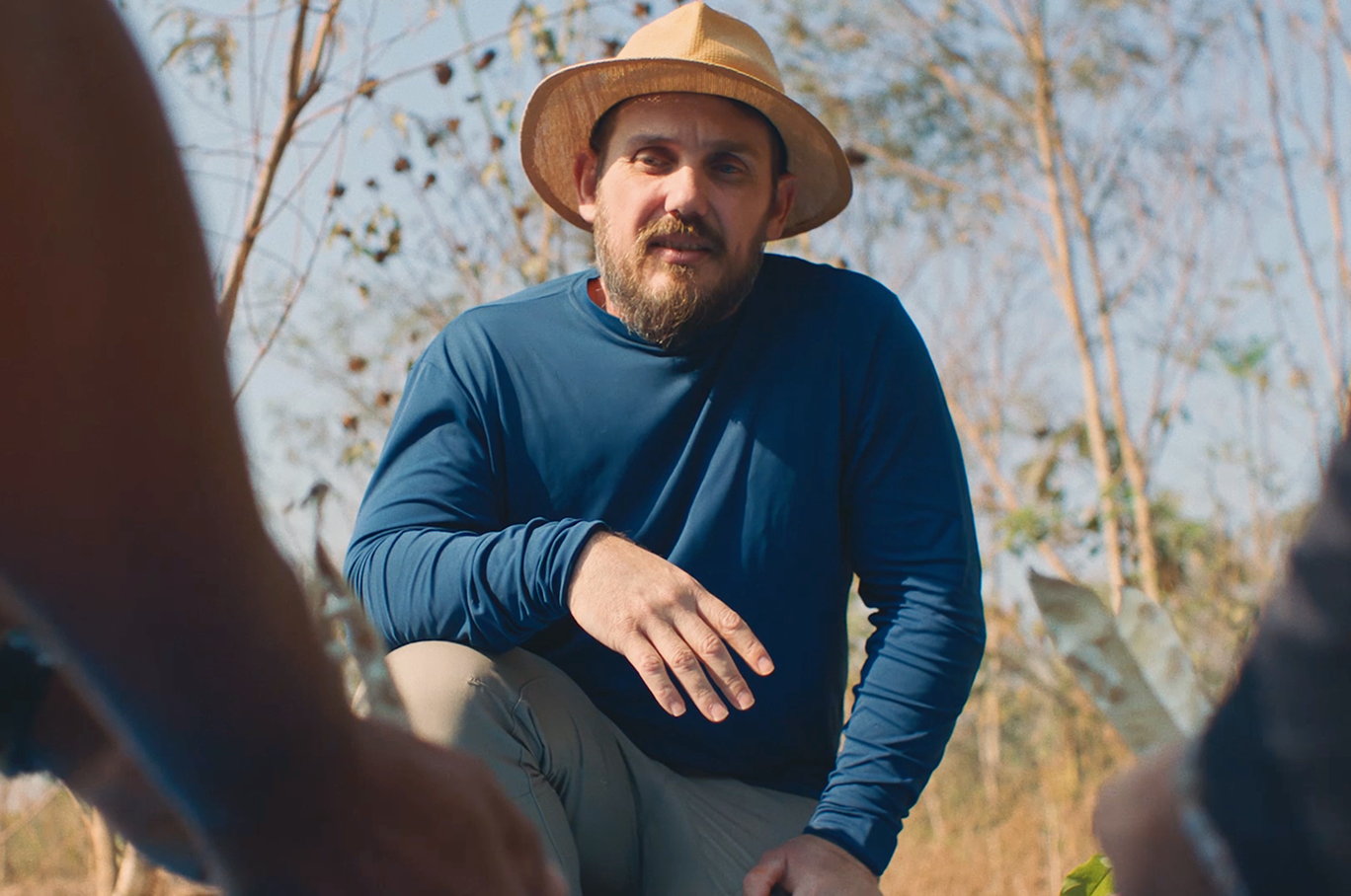 Um homem com chapéu de palha, agachado ao ar livre.