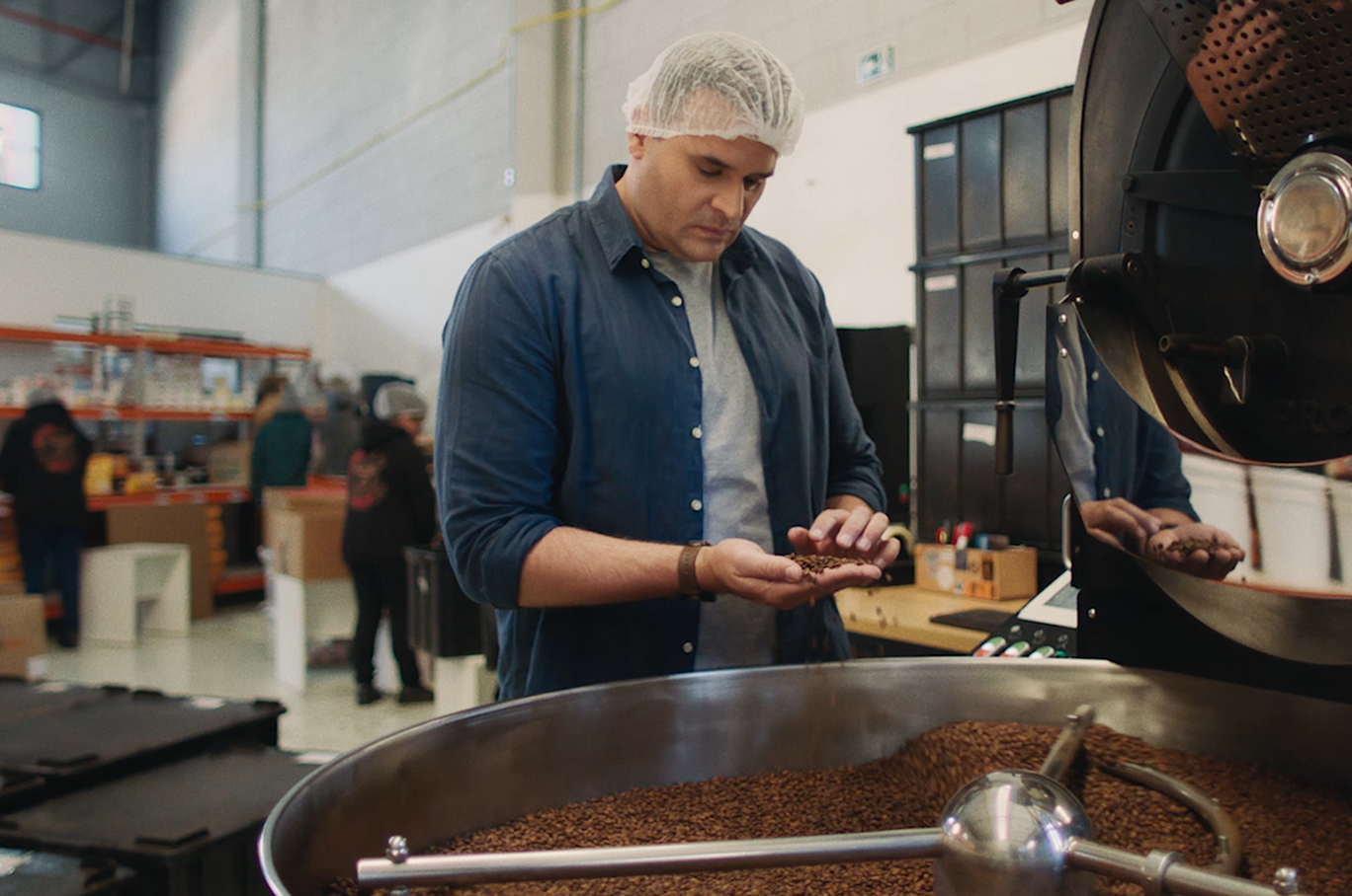 Um homem segurando grãos de café, com uma máquina de torrefação ao lado. 