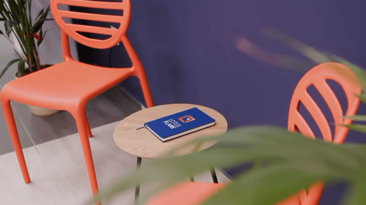 A blue notebook with the "Lotus Flare" logo sits on a small, round wooden table between two orange chairs against a purple wall.