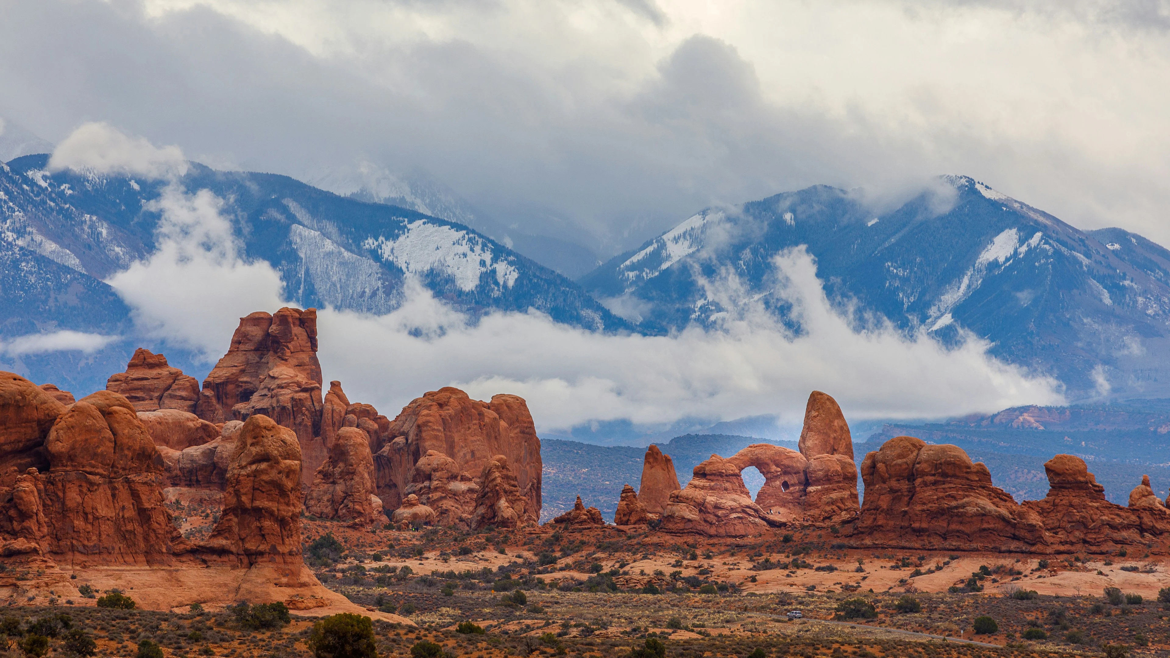 Utah desert landscape with red rock formations and distant, snow-covered mountains shrouded in low clouds.