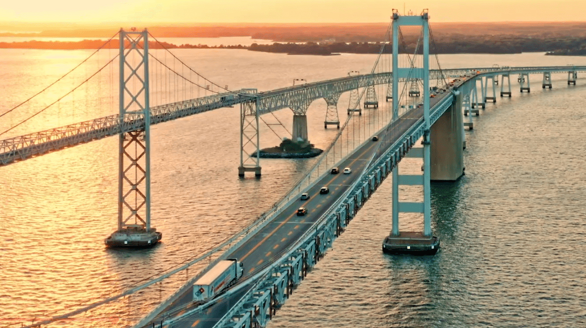 An aerial view of the Chesapeake Bay Bridge in Maryland at sunset with cars and a semi-truck driving across the water.