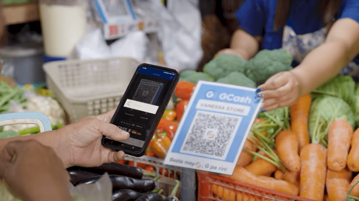 A person scanning a GCash QR code with a smartphone at a market stall filled with fresh vegetables.