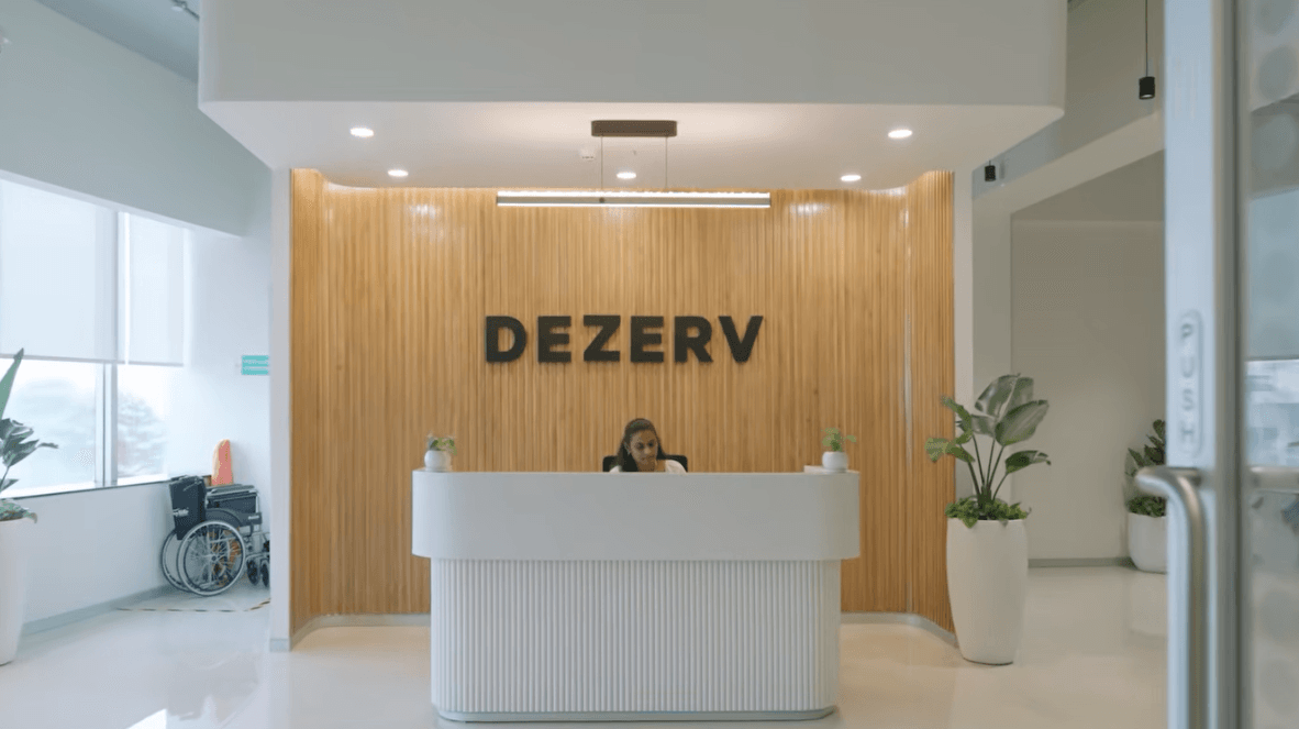 A woman sat at a white reception desk in front of a wooden slatted wall featuring the "DEZERV" logo.
